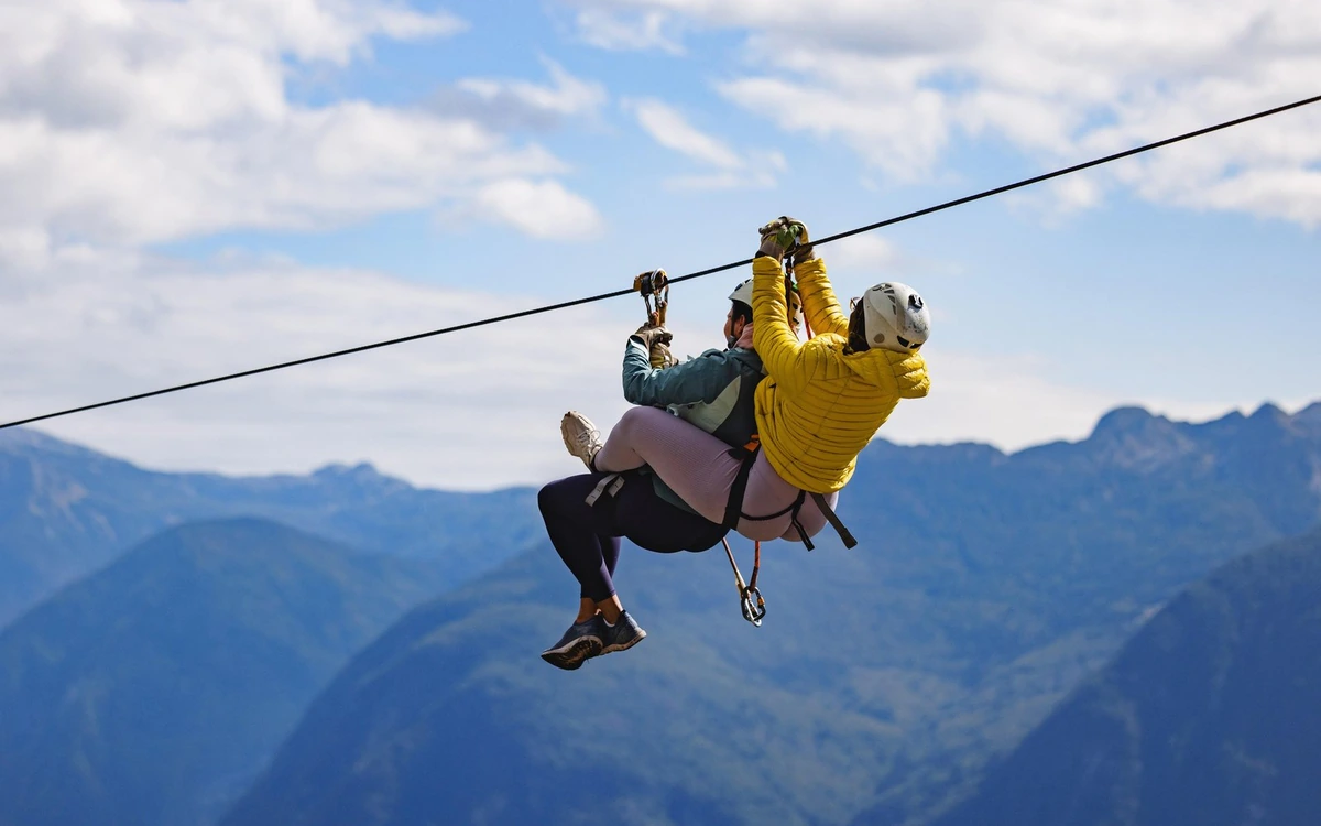 Two riders side by side on one zipline cable with mountain ridges behind them