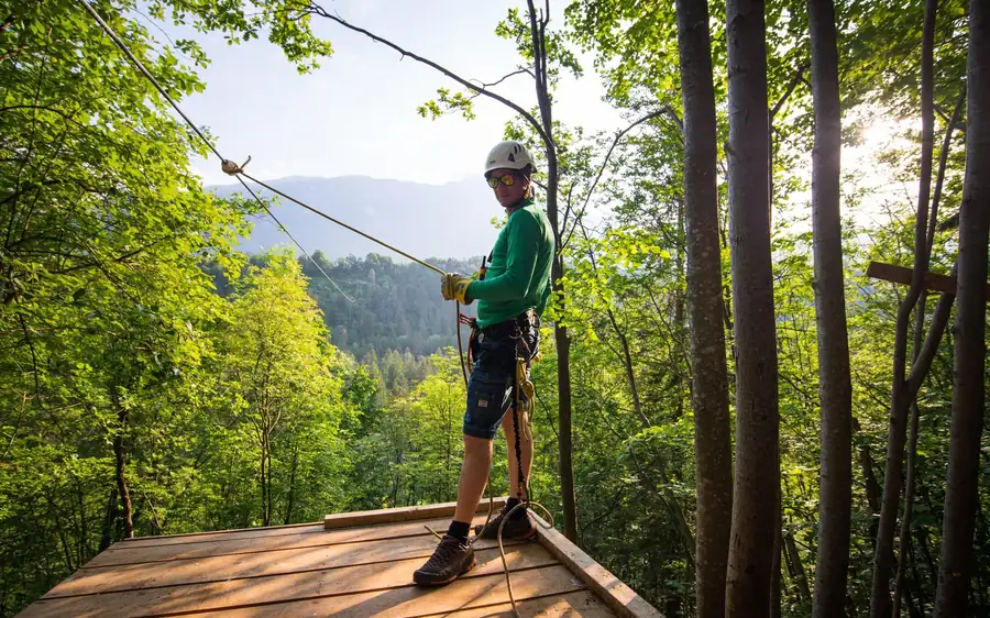 A rider clipped to the cable on a wooden launch platform among the trees, wooded hills behind