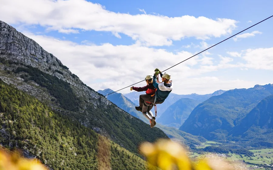 Two riders on one cable high above a broad green valley ringed by mountains